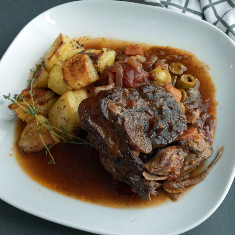 Roasted piece of lamb leg on vegetables in a red wine tomato sauce, served with roasted potatoes