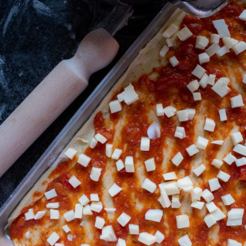 raw pizza dough flattened on a baking sheet with tomato sauce and mozzarella dices