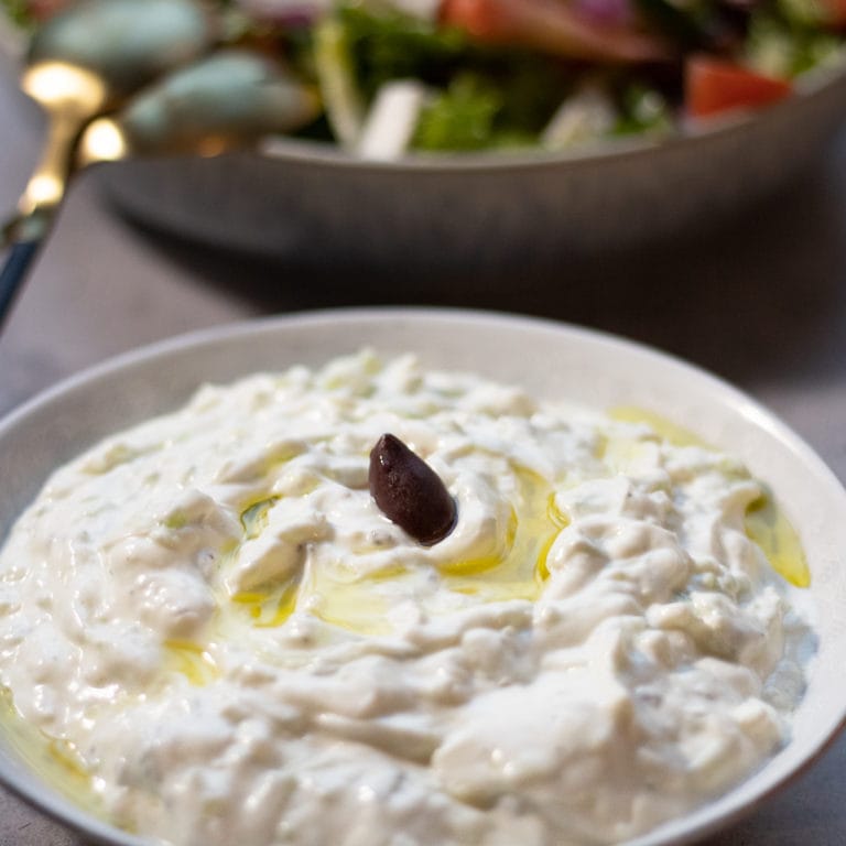 Bowl of tzatziki garnished with an olive and greek salad in the background