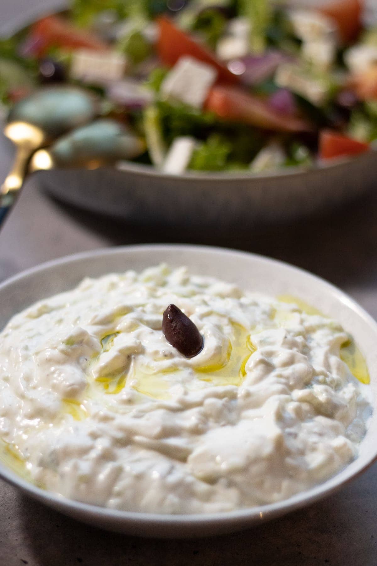 Bowl of tzatziki garnished with an olive and greek salad in the background